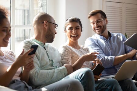 Diverse friends gathered together spend free time with modern devices sitting on sofa having fun...