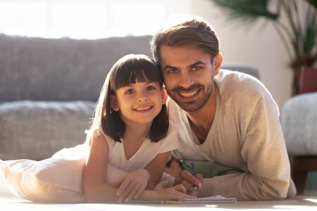 Portrait of happy young dad cuddle lying on floor entertain draw with cute preschooler daughter, smiling father and little kid look at camera have fun painting picture enjoy weekend at home togetherの写真素材