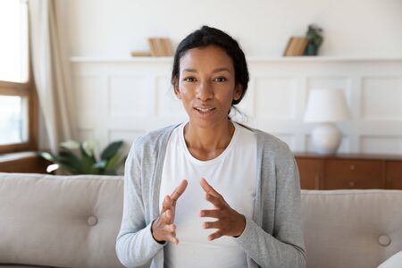 Close up headshot portrait of focused African American young woman sit on couch talk on video call,...