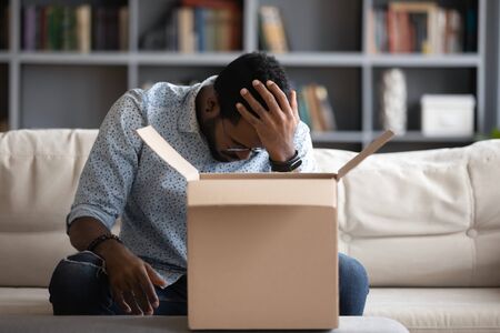 Stressed young multiracial man sitting on couch at home, feeling desperate about ordering wrong item. Depressed african american male client unpacked delivery box, dissatisfied with purchase.の写真素材
