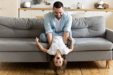 Overjoyed loving young father sit on couch in kitchen feel playful have fun with cute little daughter, happy dad play engaged in funny activity with small girl child enjoy family weekend at homeの写真素材