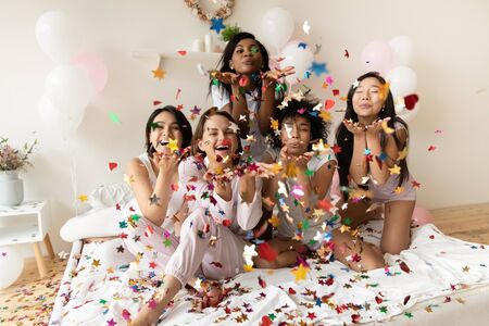 Portrait happy diverse girls wearing pajamas blowing confetti at camera, having fun at sleepover or hen party, sitting on bed in hotel room with pink balloons, five beautiful young women celebratingの写真素材