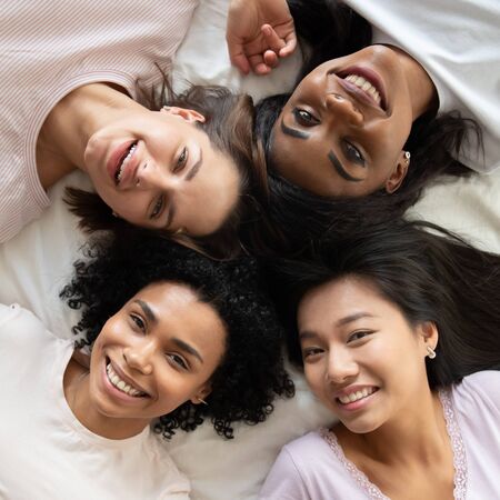 Top view diverse smiling young women best friends wearing pajamas relaxing lying in bed together, portrait from above beautiful girls looking at camera, having fun at hen-party, square photoの写真素材