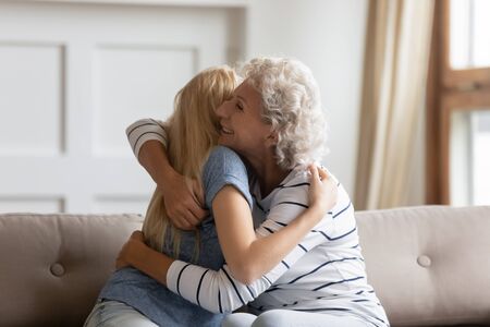Happy senior mom and grownup daughter sit on couch hug cuddle make peace after family fight, smiling...