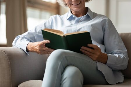 Close up of smiling senior 60s woman sit relax on couch in living room reading paper book, happy senior 70s grandmother rest on sofa ta home enjoy interesting novel or literature, hobby conceptの写真素材