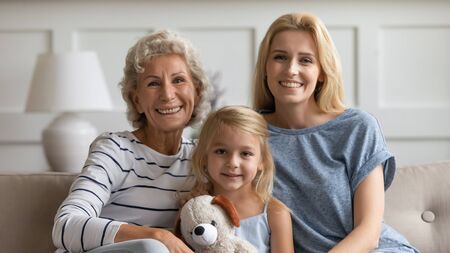 Family portrait of happy three generations of women sit relax on couch in living room look at camera posing, smiling young woman with mature mother and preschooler daughter rest on sofa at homeの写真素材