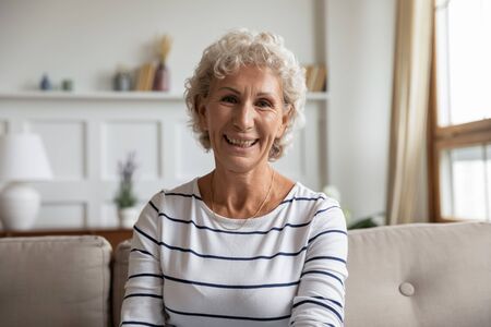 Headshot portrait of smiling senior woman sit on couch in living room having video call or...