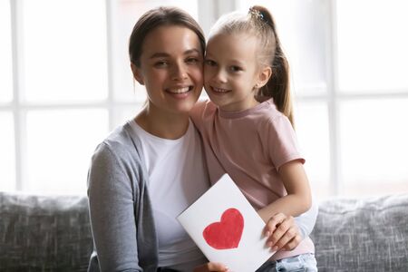 Smiling attractive young mother holding greetings card, cuddling little daughter, looking at camera. Portrait of positive two generations family sitting on sofa, celebrating birthday or special event.の写真素材
