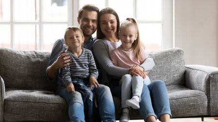 Portrait of young married couple resting on sofa, holding on lap adorable kids siblings. Happy loving bonding family of four relaxing on couch, posing for photo together in modern living room.の写真素材
