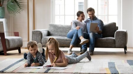 Full length small playful boy lying on floor carpet with happy elder sister, drawing pictures in...