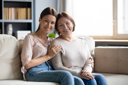 Portrait of smiling young woman cuddling affectionate older mother, sitting together on cozy sofa at...