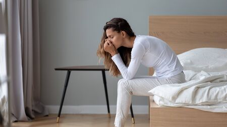 Depressed millennial girl sit on bed in home bedroom thinking pondering suffering from depression, upset unhappy young woman lost in thoughts struggle with relationship problem or psychological dramaの写真素材