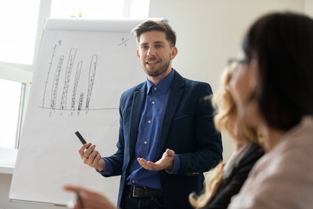 Confident millennial Caucasian male tutor coach talk present project on whiteboard, successful young businessman speak lead meeting with diverse coworkers, make flip chart presentation at briefingの写真素材