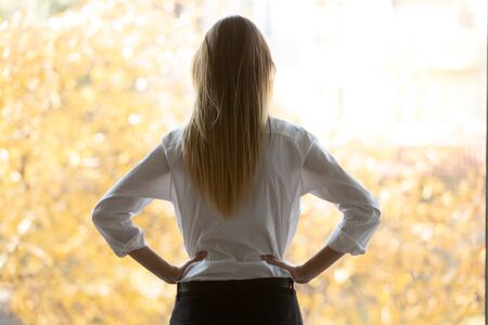 Rear view thoughtful millennial business woman looking at office window. Puzzled young female team...