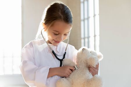 Head shot smiling adorable small girl wearing white coat, using stethoscope on teddy bear. Attractive little preschooler in uniform playing doctor patient game in clinic, children healthcare concept.の写真素材