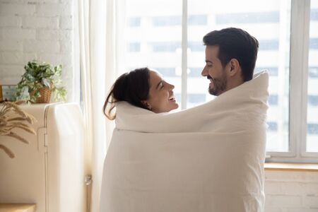 Happy loving couple wrapped in white warm blanket hugging, standing in kitchen at home, smiling beautiful wife and handsome husband looking at each other, enjoying tender moment, having funの写真素材