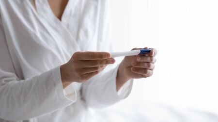 Close up focus on quick plastic pregnancy test in female hands. Young woman in white nightgown pajamas bathrobe looking at stick, waiting for results, fertility maternity check, healthcare concept.の写真素材