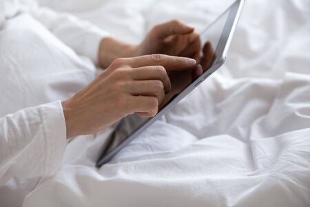 Close up young woman lying in comfortable bed under duvet, using computer digital tablet after...