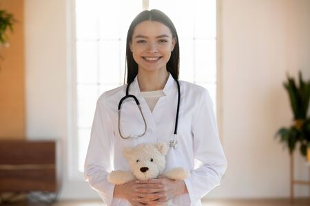 Portrait of pleasant smiling nurse medical specialist, holding toy. Happy young female general practitioner in white coat with stethoscope looking at camera, medical insurance healthcare concept.の写真素材