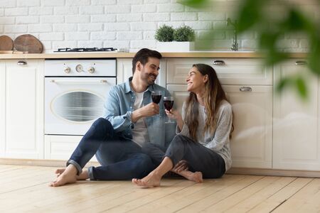 Happy loving couple clinking red wine glasses in modern kitchen, sitting on warm wooden floor together, smiling wife and husband celebrating relocation or anniversary, having fun, romantic dateの写真素材