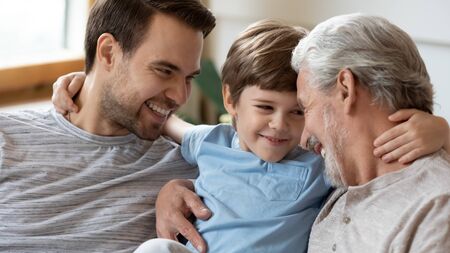 Overjoyed three generations of men hug cuddle enjoy weekend at home together, happy little preschooler boy embrace relax in living room with smiling young dad and mature grandfatherの写真素材