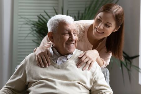 While elderly man rest sit on comfy armchair his nurse hug him showing care. Grown up granddaughter...