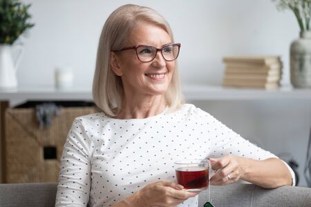 Head shot close up portrait happy stylish older lady in eyewear relaxing alone with cup of tea on comfortable sofa at home. Smiling middle aged woman enjoying weekend leisure free time, dreaming.の写真素材