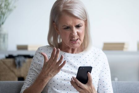 Unhappy middle aged lady irritated by bad smartphone work, head shot close up. Angry mature woman received message with bad news, annoyed by spam sms or scam email, poor internet connection.の写真素材