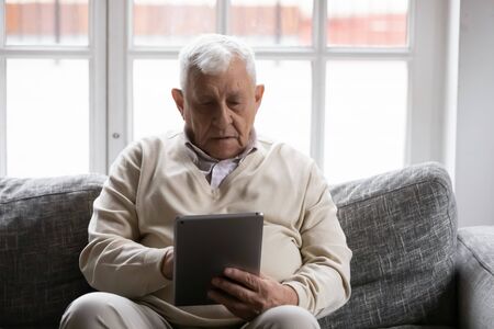 80s mature hoary man spend time seated on couch at home with tablet device, makes video call to therapist, learn helpful application. Older generation and modern technology gadgets easy usage conceptの写真素材