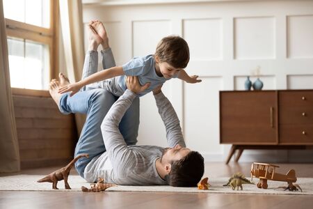 Happy young father lying on floor in living room hold fly with little preschooler son engaged in funny game together, loving dad relax playing with small boy child, enjoy family weekend at homeの写真素材
