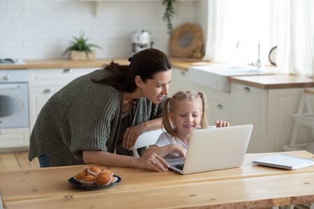 Loving young mum and little preschooler daughter sit at kitchen table using laptop together, caring mother help small girl child teaching learning online on computer, kids and technology conceptの写真素材