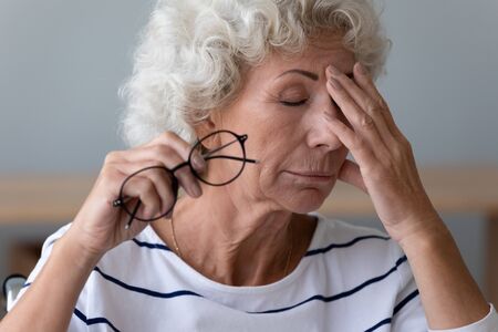 Exhausted mature woman take off glasses suffer from blurry vision or dizziness, have high blood pressure, tired senior female struggle with headache or migraine at home, elderly sight problem conceptの写真素材