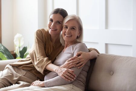 Portrait happy adult daughter and older mother hugging and holding hands, sitting on couch at home,...