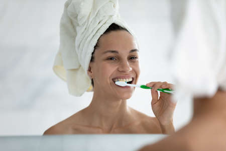 After shower 35s beautiful woman wrapped head in towel looking in mirror brushing teeth, view over female shoulder. Personal hygiene, morning routine, periodontal disease and caries prevention conceptの写真素材