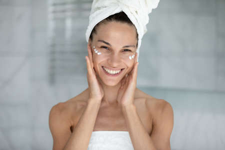 Head shot portrait perfect woman with white wide smile wrapped in white towel after shower applied day cream, touch clean ideal skin looking at camera, skincare and natural beauty treatment conceptの写真素材