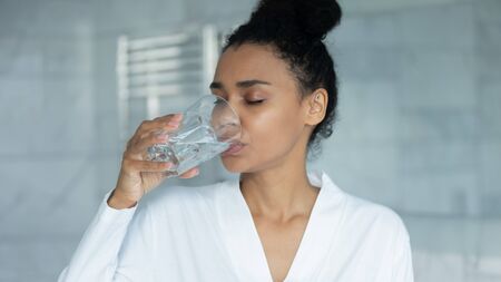 African woman in bathrobe holding glass drink filtered still clear water, preventing of skin...