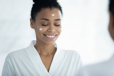 African woman with eyes closed in bathrobe reflected in mirror applied facial cream moisturizer for...