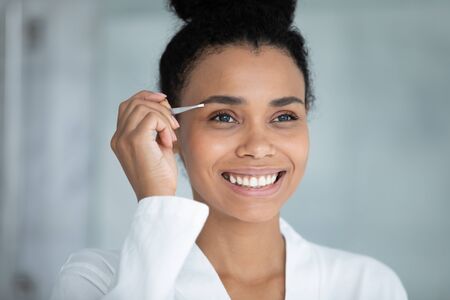 African beautiful woman with white perfect teeth charming wide smile holding metal tweezers tweezing eyebrows do correction procedure in bathroom improves shape caring about face skin beauty conceptの写真素材