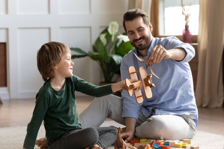 Smiling two male generations family playing with toys, sitting on floor carpet, enjoying playtime together in modern living room. Excited young man having fun with little kid boy son at weekend time.の写真素材