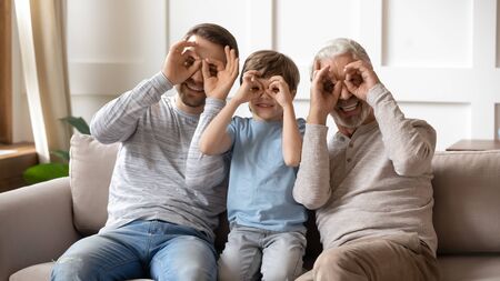 Portrait of overjoyed three generations of men sit on couch posing for funny picture together, smiling little boy with young dad and senior grandfather make face gestures look at camera at homeの写真素材