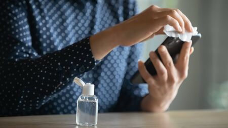 Focus on plastic bottle with antibacterial gel liquid on table with young girl cleaning smartphone screen on background. Cautious indian girl sanitizing electronic gadget, killing virus infection.の写真素材