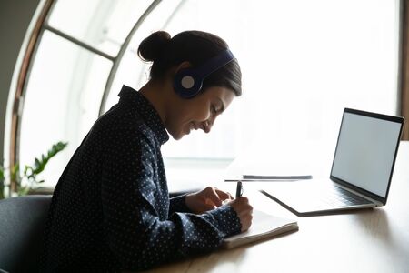Side view happy young indian woman sitting at table with laptop, wearing earphones, enjoying studying remotely from home. Smiling mixed race female student learning on online courses, writing notes.の写真素材