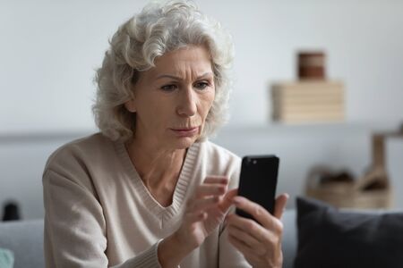 Head shot worried senior mature retired woman holding phone, reading unpleasant news in social media. Unhappy older middle aged grandmother sitting on sofa, received bad news message on smartphone.の写真素材