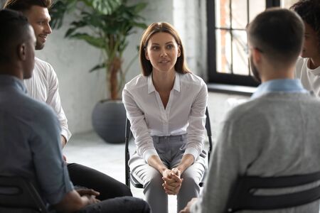 Smiling attractive female psychologist counselling speaking with diverse people sitting in circle at group therapy session. Business coach training staff, having fun, team building activity at work.の写真素材