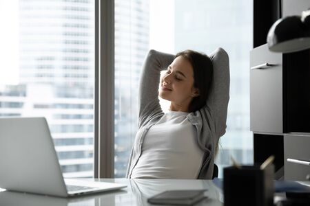 Happy young businesswoman sit at desk in office stretch in chair after finished computer work, smiling relaxed millennial female employee rest exercise at workplace, take break from jobの写真素材
