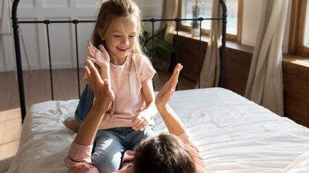 Happy little girl sitting on mommys belly, playing patty cake in bedroom. Playful small adopted child clapping hands with joyful foster parent, having fun enjoy family weekend together on bed.の写真素材