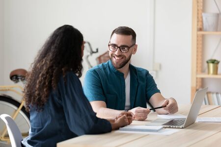 Smiling diverse businesspeople sit at desk laugh brainstorming working together on laptop at meeting, happy colleagues have fun discuss business ideas or project, cooperating on computer at briefingの写真素材