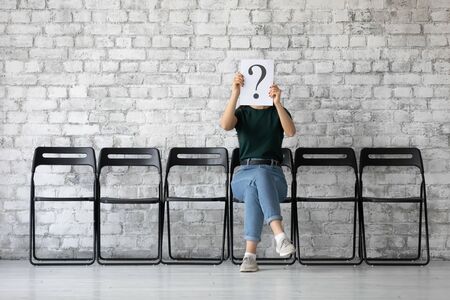 Young female job applicant sit on chair in corridor hide face hold sheet paper with question mark, millennial woman work candidate wait for interview or recruitment talk, hiring, employment conceptの写真素材