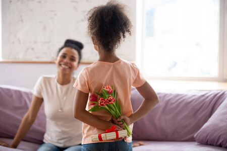 Rear view african american daughter holding flowers, bouquet and gift behind back, congratulate young diverse mother with mothers day or birthday, adorable child making surprise to mum at home.の写真素材