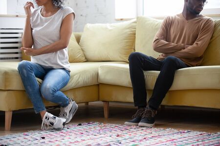 Cropped close up African American family ignoring each other, wife and husband sitting separately on sofa at home, unhappy girlfriend and boyfriend not talking after quarrel, break up or divorceの写真素材